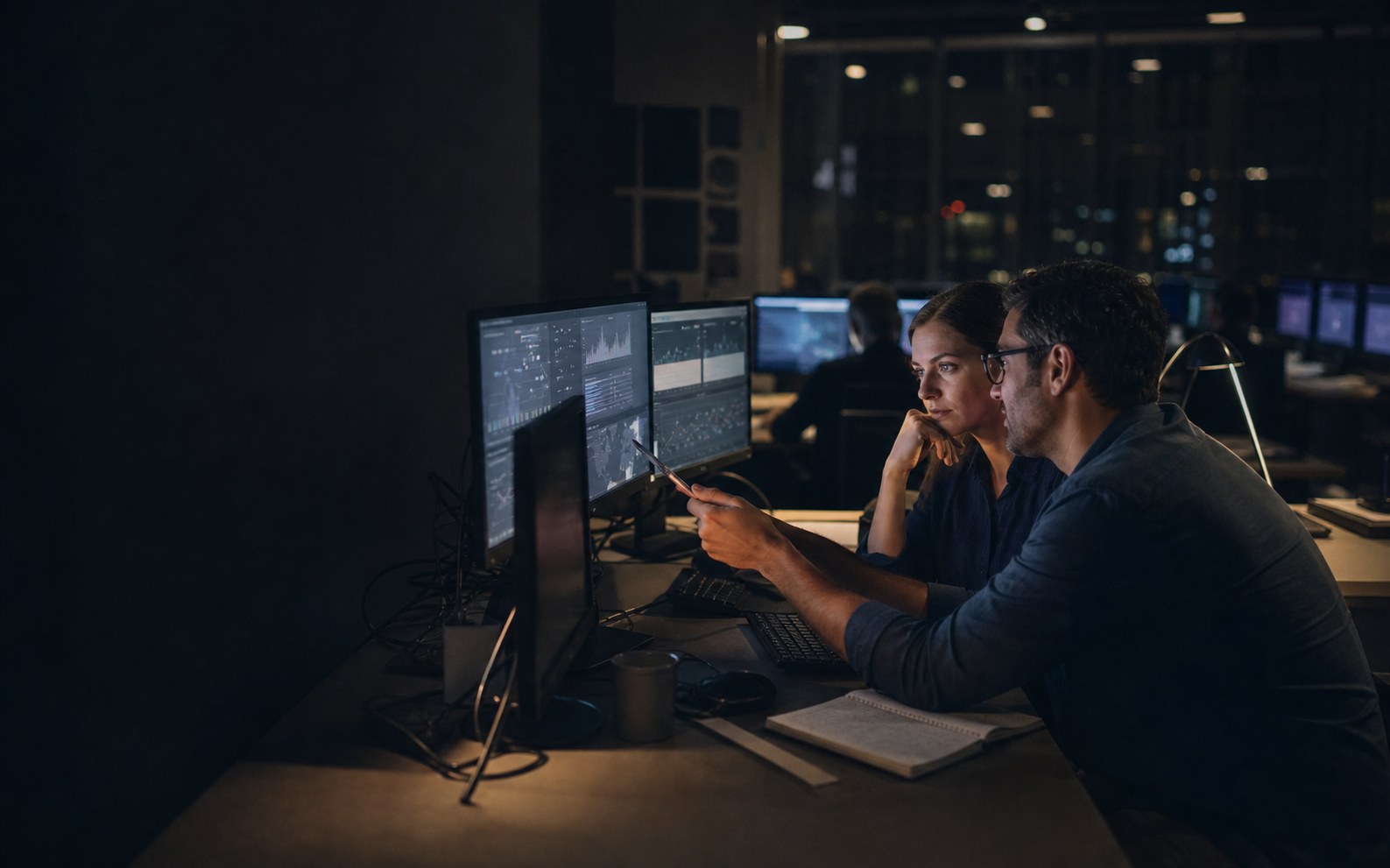 Editorial illustration of two risk analysts working at a private in-house incident desk at night.